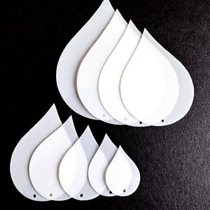 May include: A collection of white, teardrop-shaped stencils of varying sizes, arranged on a black surface. Each stencil has a small hole at the bottom. The stencils are made of a translucent material.