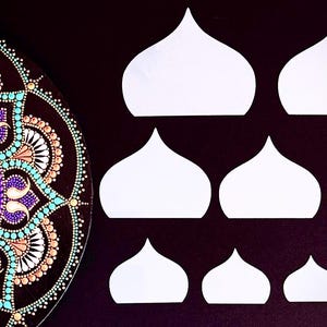 Op de afbeelding: Een gedetailleerd mandala-ontwerp in tinten teal, paars en perzik op een zwarte achtergrond. Verschillende witte papieren uitsparingen in een koepelvorm zijn op een donkere ondergrond gerangschikt, variërend in grootte.