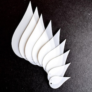 May include: White, teardrop-shaped plastic cutouts arranged on a black background. The cutouts are layered, creating a visual effect. Each cutout has a small hole near the pointed end. The shapes vary slightly in size.
