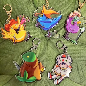 May include: Five colorful keychains featuring cartoon characters. The keychains are made of clear acrylic and feature a variety of characters, including a yellow bird with a red hat, a blue bird with a yellow hat, a purple bird with a crown, a green creature with a brown cloak, and a white creature with a red hat and a red and white striped shirt.
