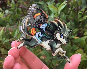 May include: A clear acrylic keychain featuring a cartoon illustration of a wolf and a green-skinned character with a mask and orange hair. The wolf is running and the character is riding on its back.