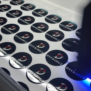 May include: A sheet of white paper with multiple black and silver round stickers. Each sticker has a red and white logo with the text "DTFKINGZ" printed on it.
