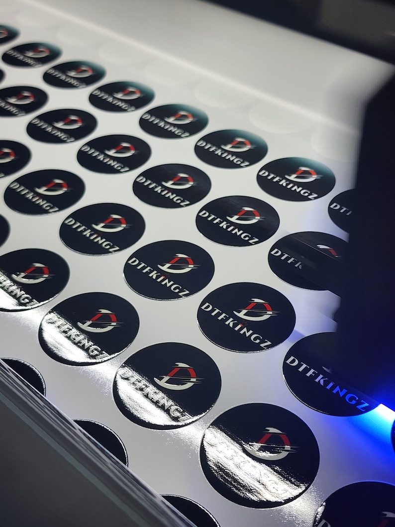 May include: A sheet of white paper with multiple black and silver round stickers. Each sticker has a red and white logo with the text "DTFKINGZ" printed on it.