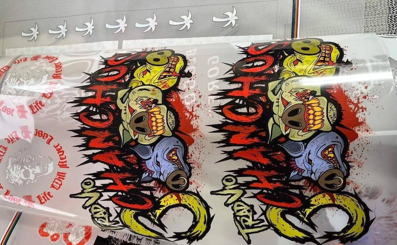 May include: A white vinyl sticker with a graphic design of three cartoon pigs with red, yellow, and blue colors. The pigs have sharp teeth and are surrounded by red and black flames. The text "TRAD NO" is written in black and red letters on the sticker.
