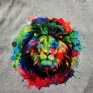 May include: A gray t-shirt featuring a vibrant, colorful lion graphic. The lion's mane and face are rendered in a watercolor style, with splashes of red, blue, green, and yellow. The design is centered on the shirt.