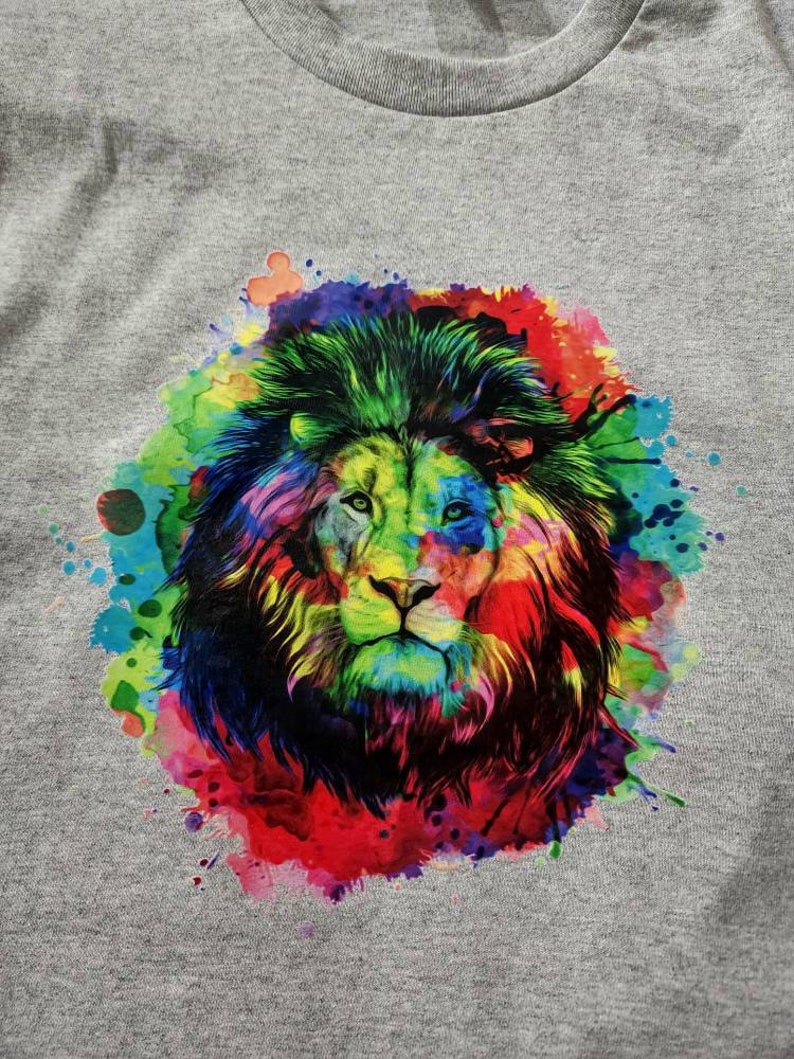 May include: A gray t-shirt featuring a vibrant, colorful lion graphic. The lion's mane and face are rendered in a watercolor style, with splashes of red, blue, green, and yellow. The design is centered on the shirt.