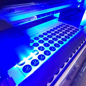 May include: A close-up of a printing machine with a sheet of round stickers being printed. The stickers are black and white and have the text "DTFLINGZ" printed on them.