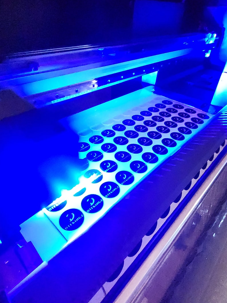May include: A close-up of a printing machine with a sheet of round stickers being printed. The stickers are black and white and have the text "DTFLINGZ" printed on them.