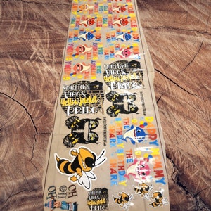 May include: A sheet of various stickers featuring cartoon sharks, bees, and text. The stickers include phrases like "Happy Birthday," "Daddy Mommy," and "Smalltown Vibes & Yellowjacket Pride." The color palette includes yellow, blue, and pink.