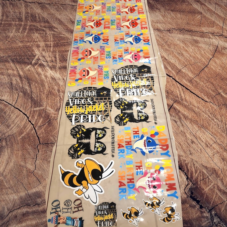 May include: A sheet of various stickers featuring cartoon sharks, bees, and text. The stickers include phrases like "Happy Birthday," "Daddy Mommy," and "Smalltown Vibes & Yellowjacket Pride." The color palette includes yellow, blue, and pink.