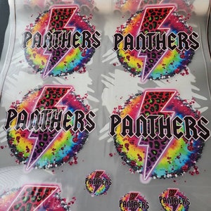 May include: A sheet of printed transfers featuring the word "PANTHERS" in a bold, black font, with a leopard print lightning bolt design. The background is a vibrant tie-dye pattern with rainbow colors. Smaller circular versions are also visible.