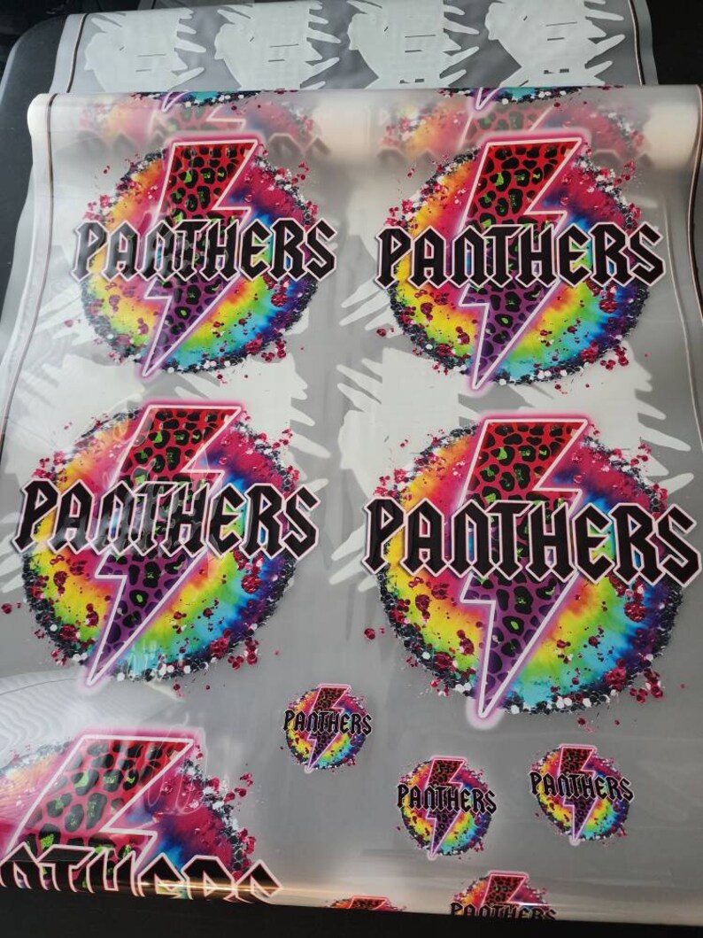 May include: A sheet of printed transfers featuring the word "PANTHERS" in a bold, black font, with a leopard print lightning bolt design. The background is a vibrant tie-dye pattern with rainbow colors. Smaller circular versions are also visible.