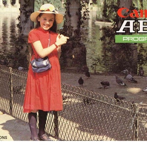 Op de afbeelding: Een jong meisje in een rode jurk en een strohoed staat voor een hek van gaas. Ze houdt een blauwe handtas vast en glimlacht naar de camera. De achtergrond is een park met een vijver en bomen. De tekst "Canon AE-1 PROGRAM" is zichtbaar in de rechterbovenhoek van de afbeelding.