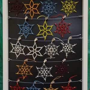 Può includere: Un'esposizione di 16 decorazioni a forma di fiocco di neve all'uncinetto in vari colori, tra cui rosso, blu, giallo e bianco. I fiocchi di neve sono appesi a una lavagna nera con cornice bianca.