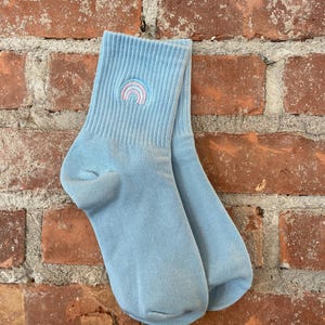 May include: A pair of pale blue socks with a ribbed cuff and a small embroidered rainbow design in pink, blue, and yellow. The socks are displayed against a red brick wall.