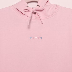 Pode incluir: Um moletom rosa com uma fileira de cinco corações bordados em azul, rosa e branco.