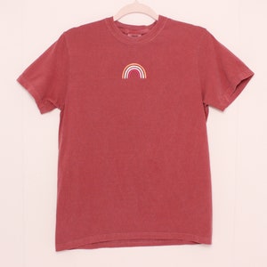 May include: A red short-sleeved t-shirt with a small embroidered rainbow in yellow, orange, pink, and white.