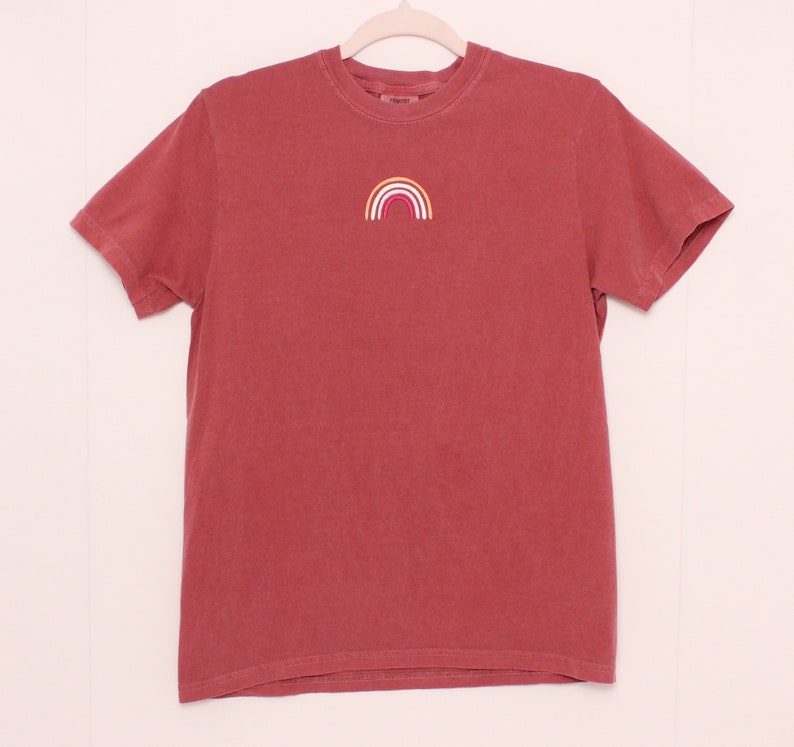 May include: A red short-sleeved t-shirt with a small embroidered rainbow in yellow, orange, pink, and white.