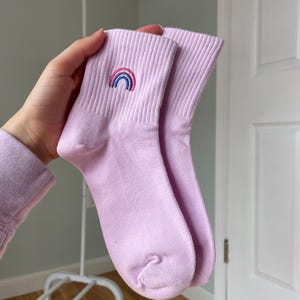 Puede incluir: Un par de calcetines de tobillo lavanda con un arcoíris bordado en un calcetín.