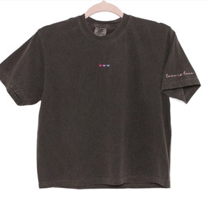 Pode incluir: Uma camiseta preta com um pequeno desenho bordado de três corações em rosa, azul e vermelho na frente. O texto "Love is Love" está bordado em rosa na manga direita.