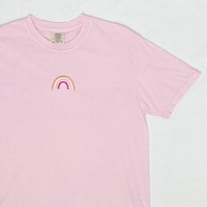 May include: A pink t-shirt with a small embroidered rainbow in orange, pink, white, and red colours. The rainbow is centred on the shirt.