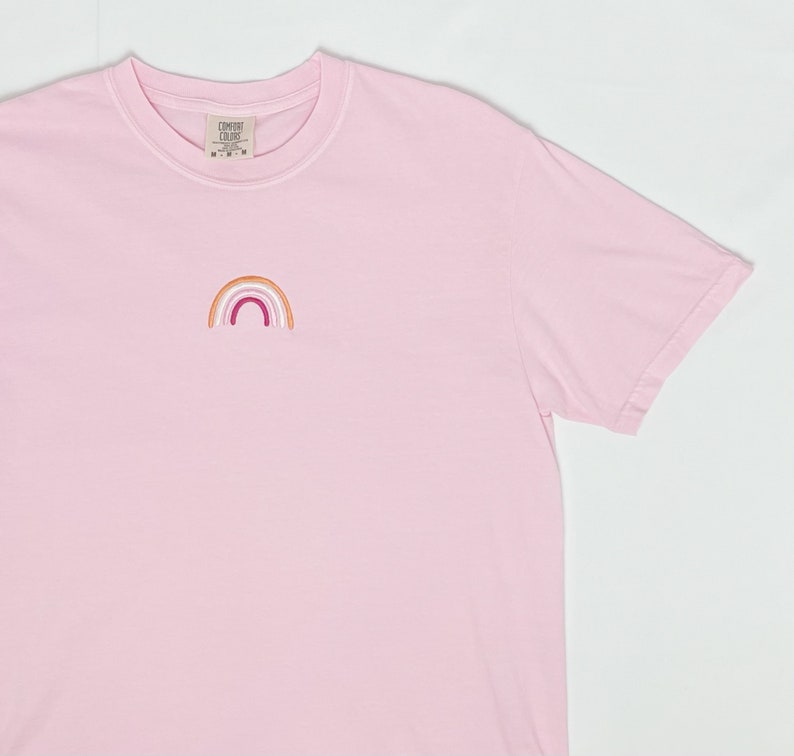 May include: A pink t-shirt with a small embroidered rainbow in orange, pink, white, and red colours. The rainbow is centred on the shirt.