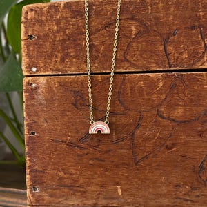 May include: A gold-colored necklace with a small rainbow pendant. The rainbow features layers of pink, white, and red. The necklace is displayed on a wooden surface with a natural wood grain.