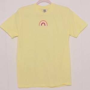 May include: A yellow short-sleeved t-shirt with a small embroidered rainbow in the centre of the chest. The rainbow is made of red, orange, yellow, green, blue, indigo, and violet.