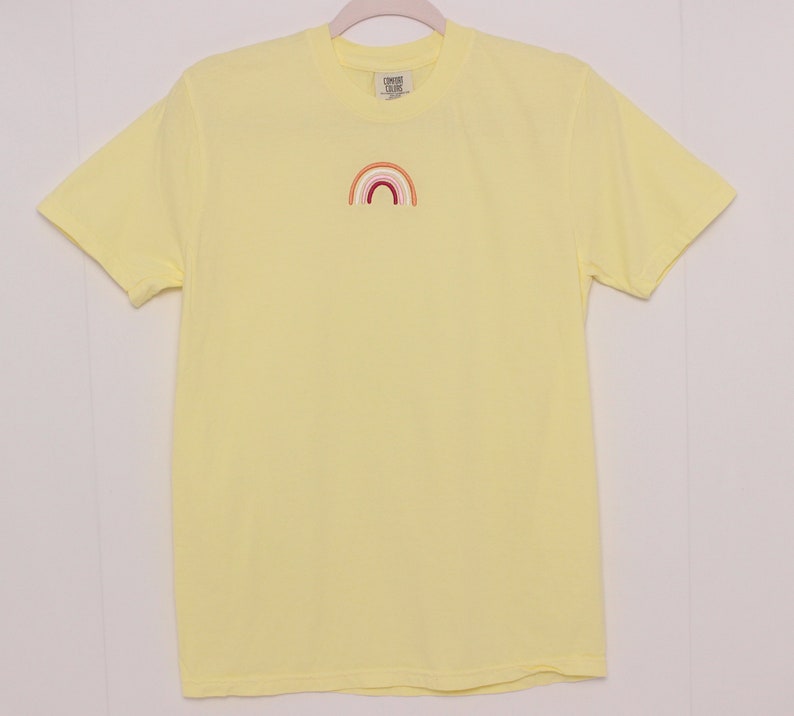 May include: A yellow short-sleeved t-shirt with a small embroidered rainbow in the centre of the chest. The rainbow is made of red, orange, yellow, green, blue, indigo, and violet.