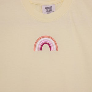 May include: A yellow t-shirt featuring an embroidered rainbow in shades of pink, orange, and white. The rainbow is centrally positioned on the shirt and has a simple, minimalist design.