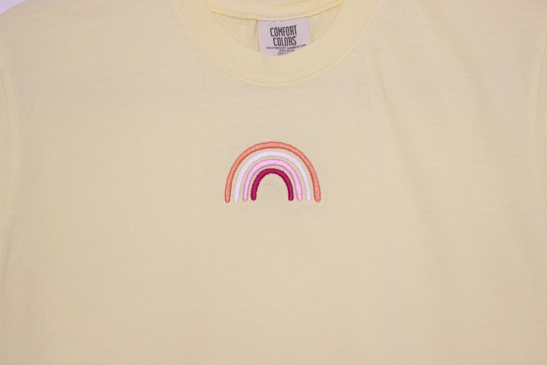 May include: A yellow t-shirt featuring an embroidered rainbow in shades of pink, orange, and white. The rainbow is centrally positioned on the shirt and has a simple, minimalist design.