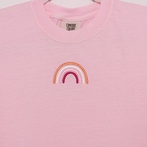 May include: A pink t-shirt with an embroidered rainbow in shades of orange, pink, white, and red. The shirt has the text "COMFORT COLORS" on the tag.