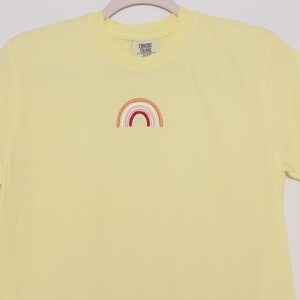 May include: A yellow t-shirt with a small embroidered rainbow in shades of orange, pink, and red.