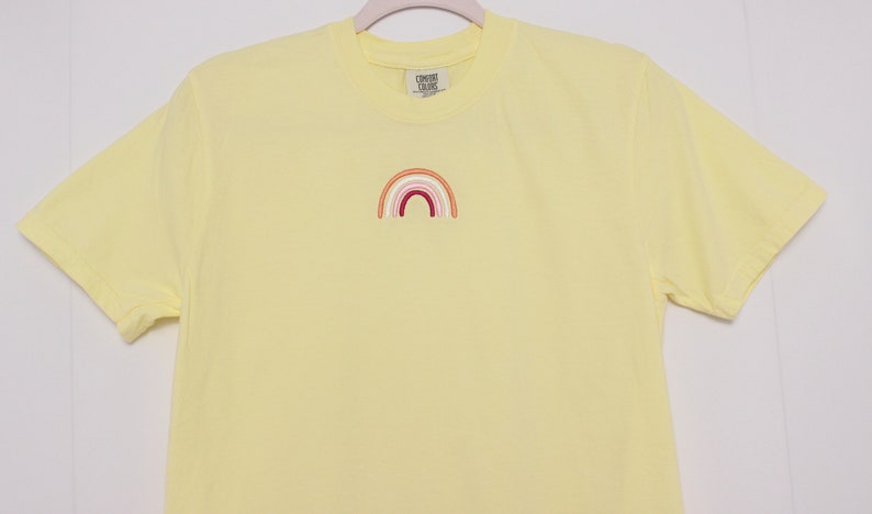 May include: A yellow t-shirt with a small embroidered rainbow in shades of orange, pink, and red.