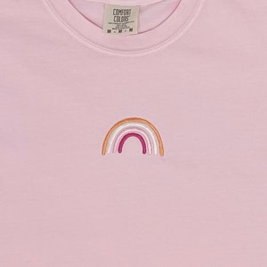 May include: A pink t-shirt with an embroidered rainbow in shades of orange, pink, and white. The shirt has a tag that reads "Comfort Colors" and "M - M - M".