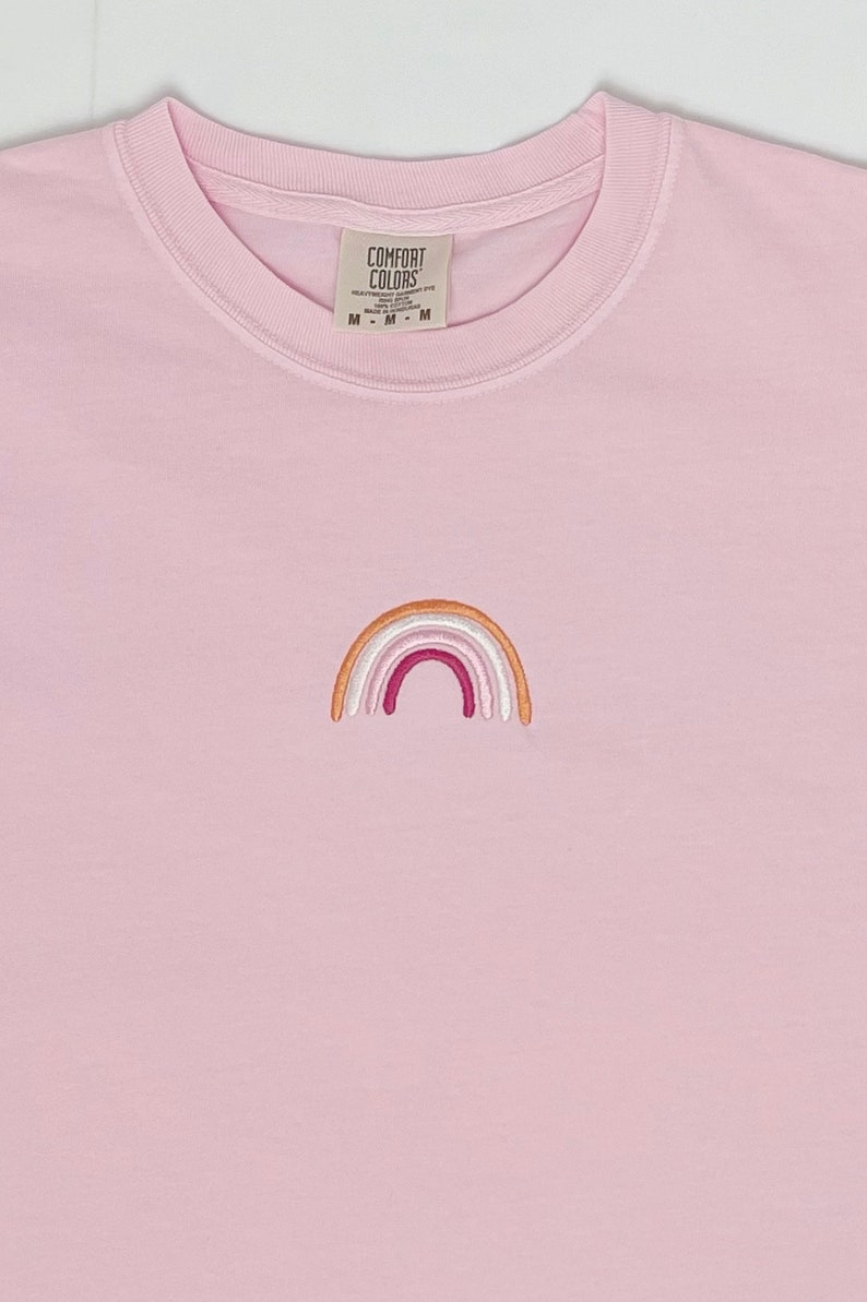 May include: A pink t-shirt with an embroidered rainbow in shades of orange, pink, and white. The shirt has a tag that reads "Comfort Colors" and "M - M - M".