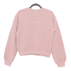 May include: A dusty rose colored sweatshirt with the text "Love is Love" embroidered in white on the front. The sweatshirt has a crew neck and long sleeves. There are small rainbow hearts embroidered on the left cuff.