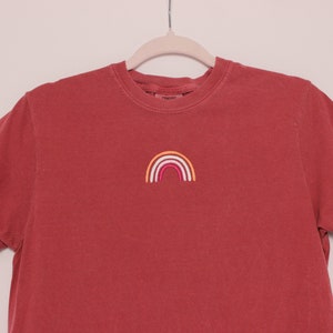May include: A red t-shirt with a small embroidered rainbow in orange, yellow, white, pink, and red.