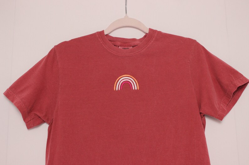 May include: A red t-shirt with a small embroidered rainbow in orange, yellow, white, pink, and red.