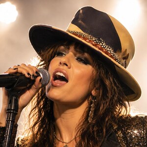 May include: A performer wearing a tan and black fedora with a feather band, a gold sequined blazer with black lapels, and a black shimmering top. The performer is singing into a black microphone while wearing layered star-shaped pendant necklaces.