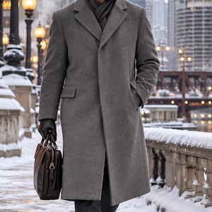 May include: A long grey wool overcoat with notched lapels, worn over dark trousers and a scarf. The outfit includes black leather gloves and a dark brown leather briefcase with gold-tone hardware, held in one hand, and black leather dress shoes.
