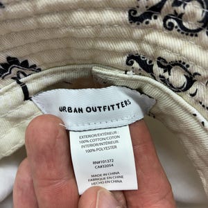 May include: Cream-colored bucket hat with a black paisley pattern. The hat has a tag that reads "Urban Outfitters" and lists the materials as 100% cotton exterior and 100% polyester interior. The tag also indicates the hat was made in China.