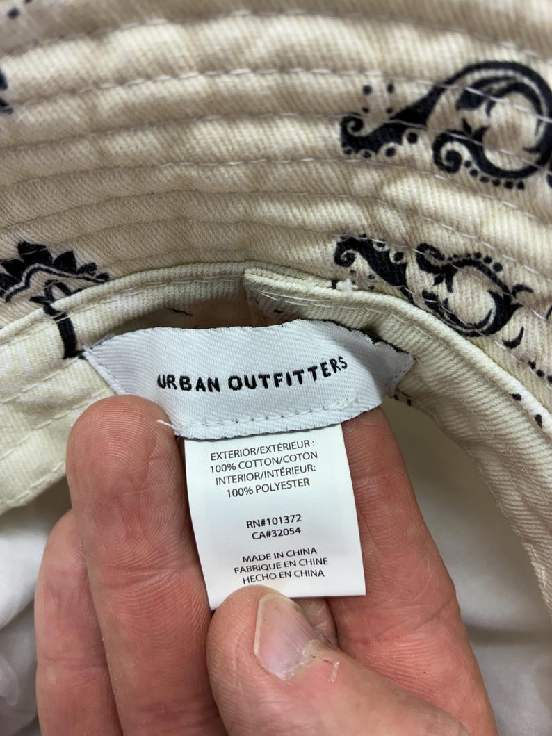 May include: Cream-colored bucket hat with a black paisley pattern. The hat has a tag that reads "Urban Outfitters" and lists the materials as 100% cotton exterior and 100% polyester interior. The tag also indicates the hat was made in China.