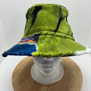 May include: A green, fuzzy bucket hat with a colorful, abstract design. The hat features a wide brim and a soft, textured material. The design includes blue, purple, and yellow accents. The hat is displayed on a white mannequin head.