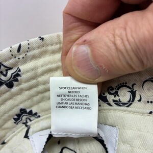 May include: Cream-colored bucket hat with a paisley pattern in black. The hat has a tag that reads "SPOT CLEAN WHEN NEEDED, NETTOYER LES TACHES EN CAS DE BESOIN, LIMPIAR LAS MANCHAS CUANDO SEA NECESARIO."