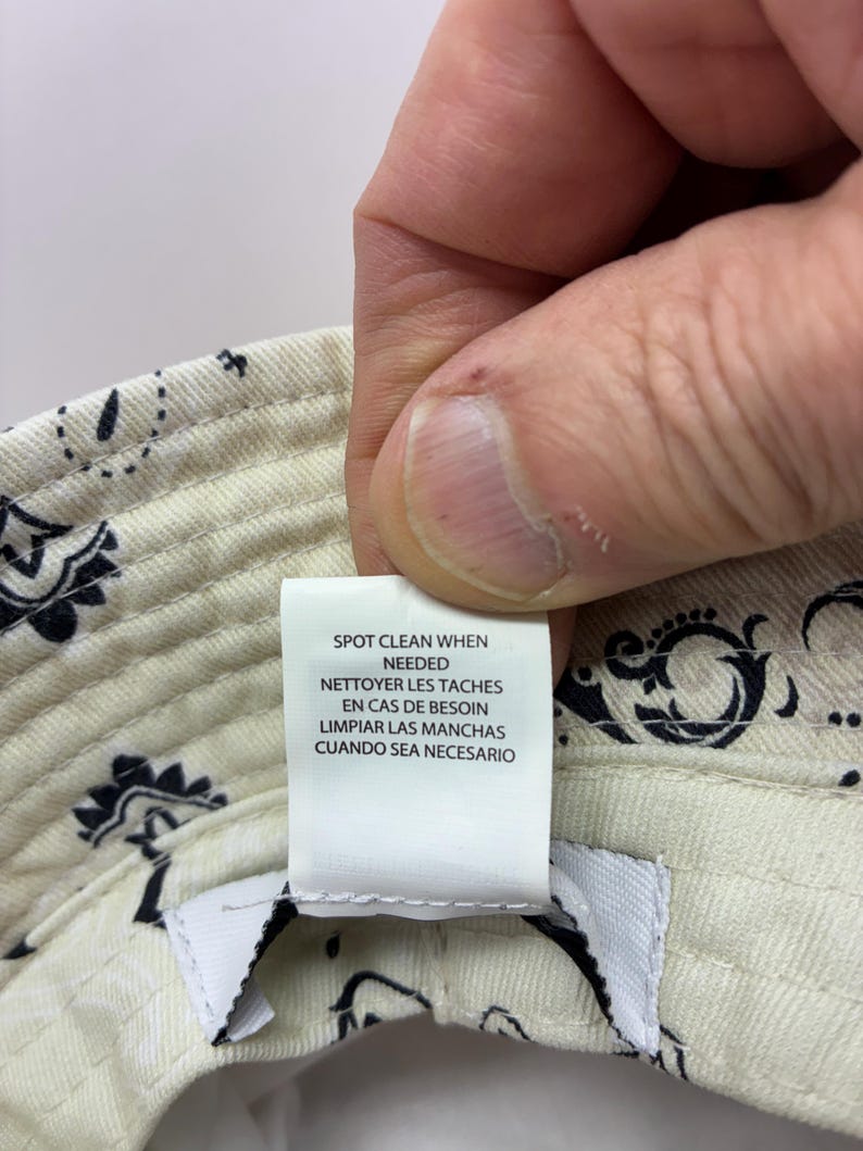 May include: Cream-colored bucket hat with a paisley pattern in black. The hat has a tag that reads "SPOT CLEAN WHEN NEEDED, NETTOYER LES TACHES EN CAS DE BESOIN, LIMPIAR LAS MANCHAS CUANDO SEA NECESARIO."
