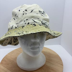 May include: Reversible bucket hat with a cream-colored exterior featuring black abstract shapes and a light green patterned interior. The hat is displayed on a white mannequin head.