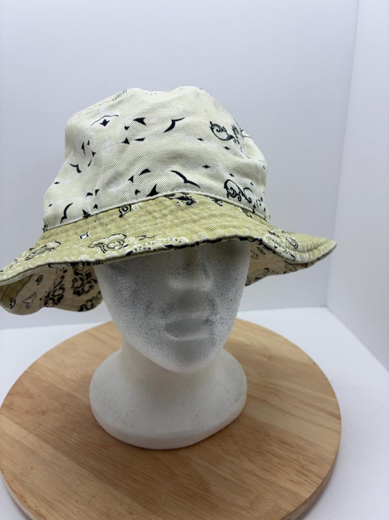 May include: Reversible bucket hat with a cream-colored exterior featuring black abstract shapes and a light green patterned interior. The hat is displayed on a white mannequin head.