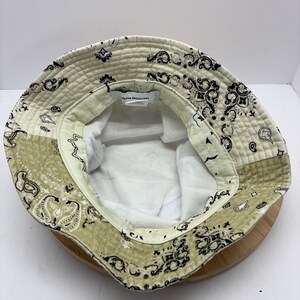May include: A reversible bucket hat with a paisley pattern in cream, olive green, and black. The hat has a white interior and the text "Urban Outfitters" is visible on the inside. The hat is sitting on a wooden surface.