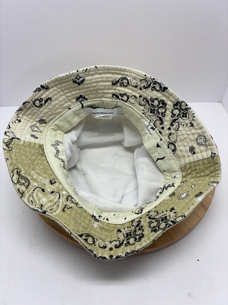 May include: A reversible bucket hat with a paisley pattern in cream, olive green, and black. The hat has a white interior and the text "Urban Outfitters" is visible on the inside. The hat is sitting on a wooden surface.